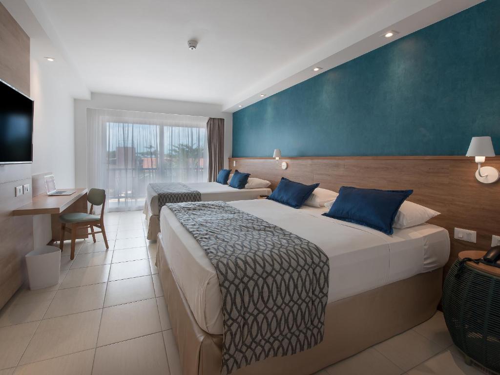 Quarto amplo com piso frio e duas camas de casal extra grande. Ao fundo, varanda com cortinas leves.
