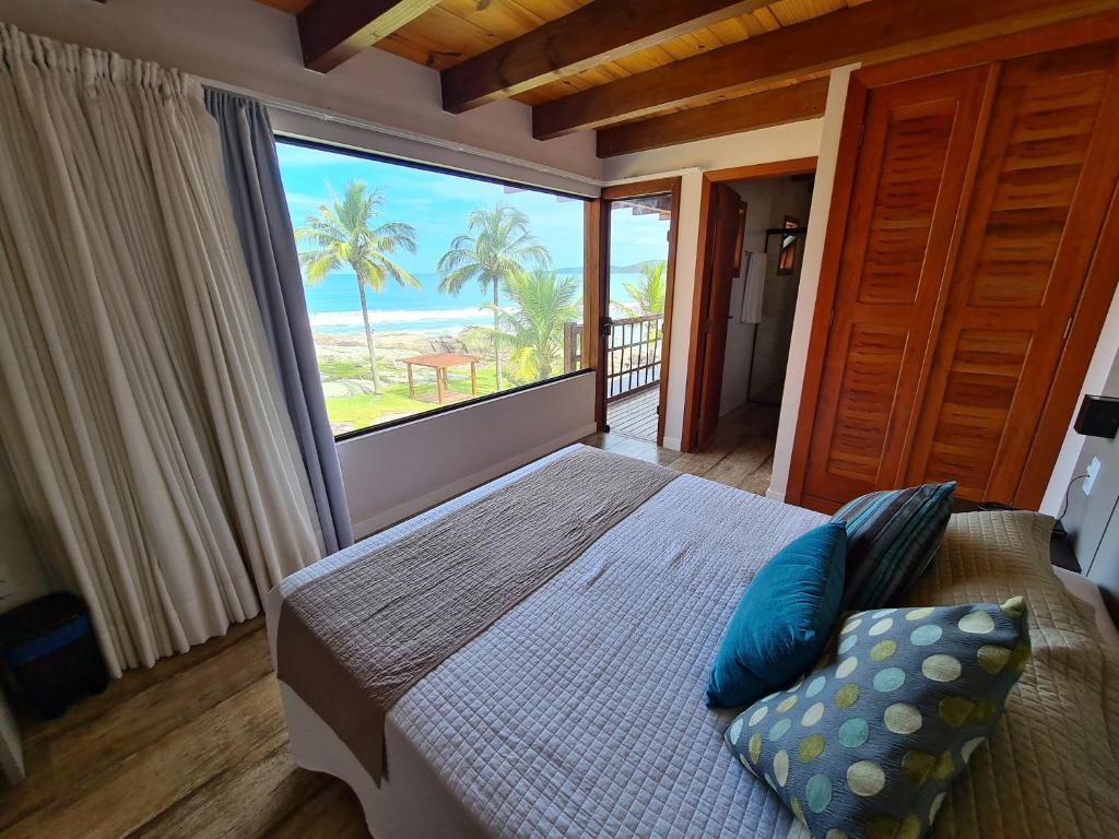 Quarto de casal com uma ampla janela de vidro que dá vista para o mar. Portinha com saída para a varanda. Pousadas pé na areia em Bombinhas.