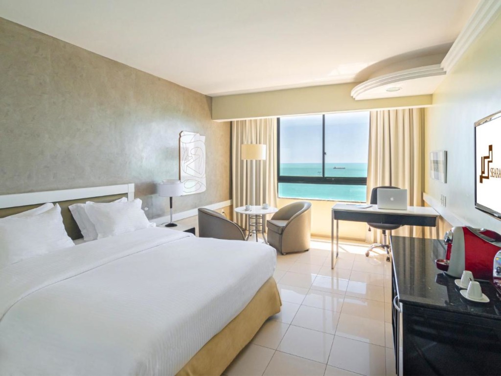 Quarto com cama de casal, máquina de café, mesa de escritório e mesinha para refeições. Ampla janela com vista para o mar em hotéis na praia do Futuro.