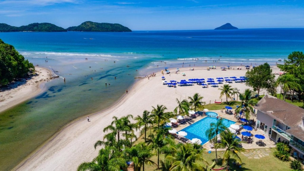 Vista panorâmica de hotel pé na areia com piscina e guarda-sóis.