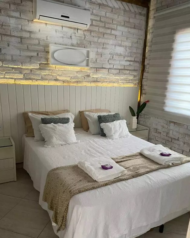 Quarto com paredes em tijolinhos brancos, cama de casal, ar-condicionado e cortina de persiana.