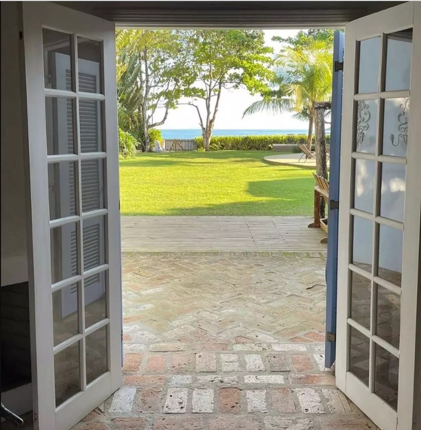 Porta balcão aberta com vista para o jardim com piscina e, ao fundo, vista do mar.